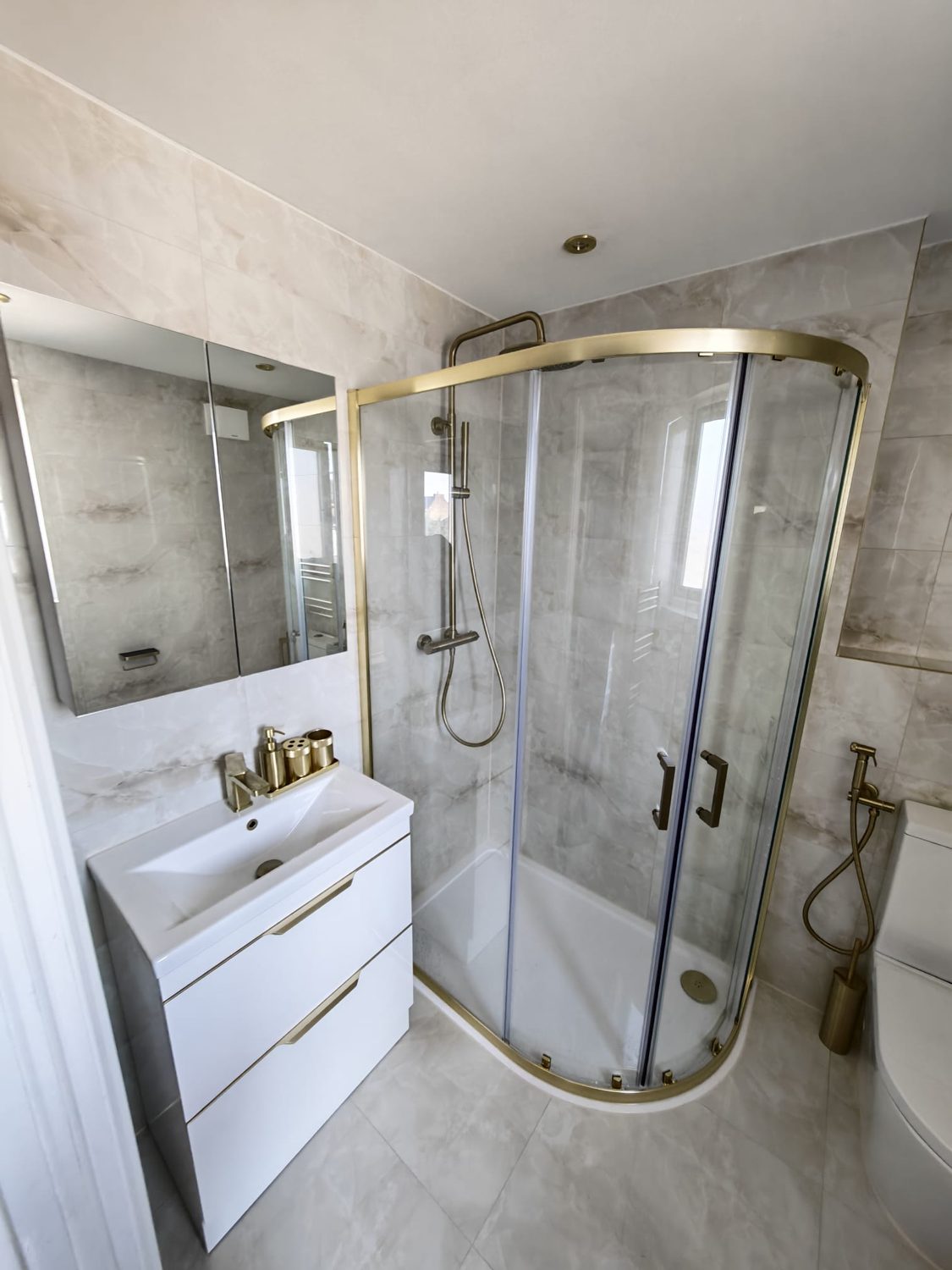 The same room finished, tiled in marble-effect porcelain with a brushed-brass quadrant enclosure and a wall-hung vanity