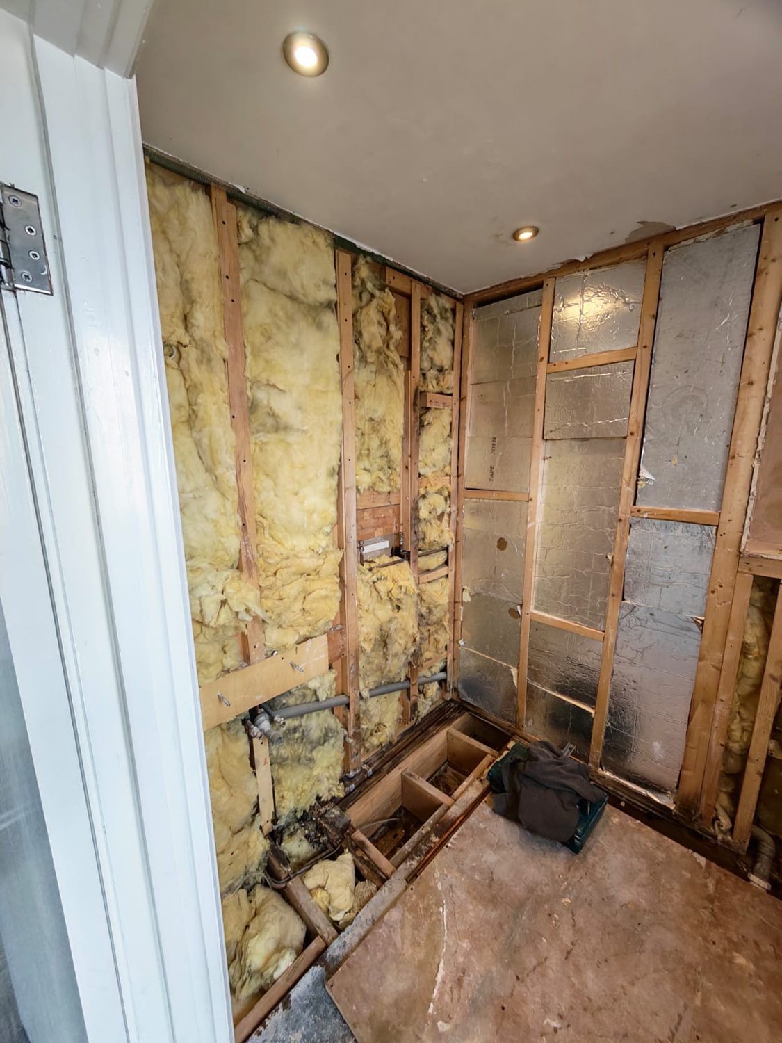 The same room opened further, showing mineral wool between the studs, foil-faced board and old pipework in the floor void