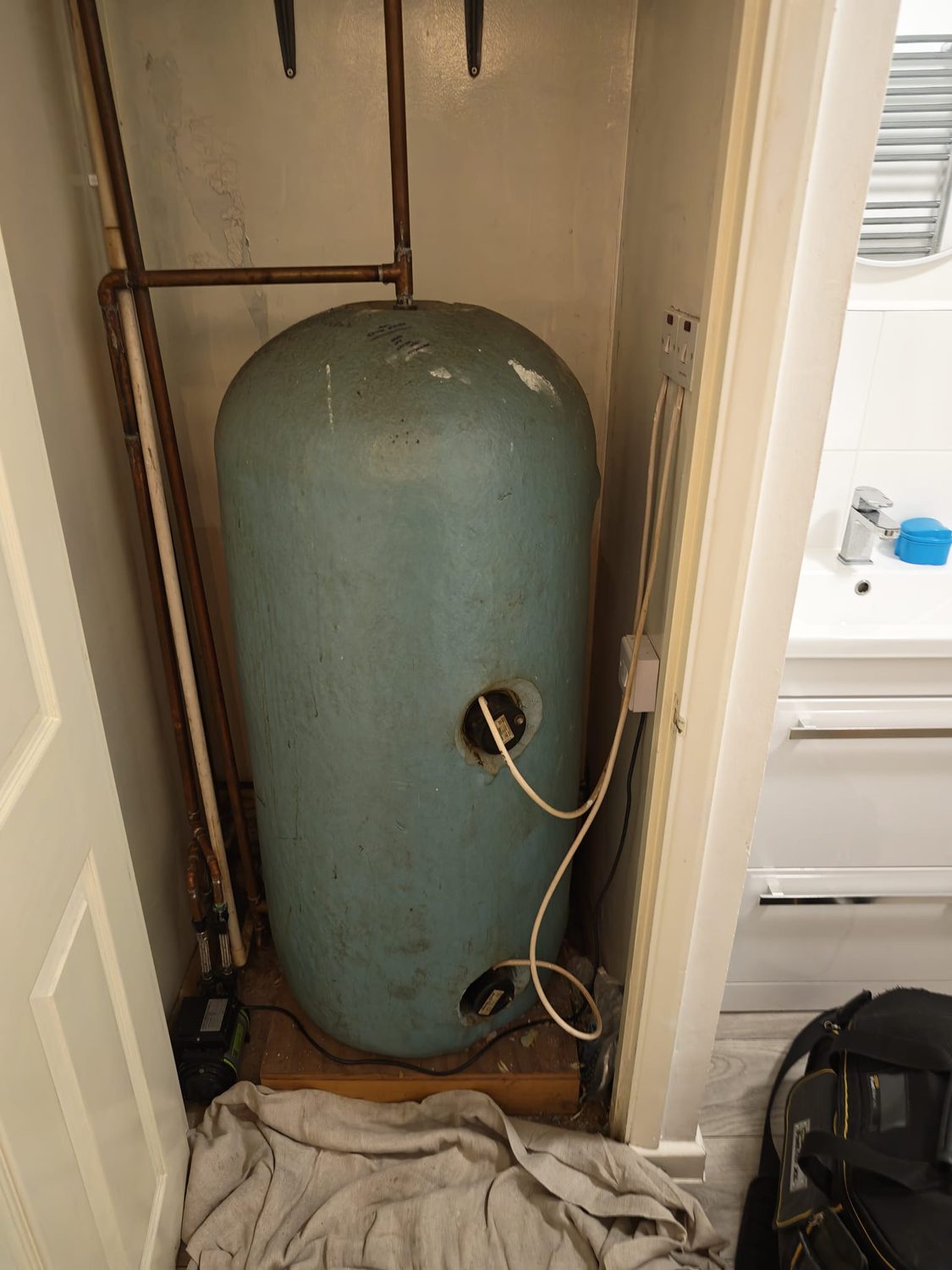 Old green-painted copper hot water cylinder in a cramped airing cupboard with corroded pipework around it