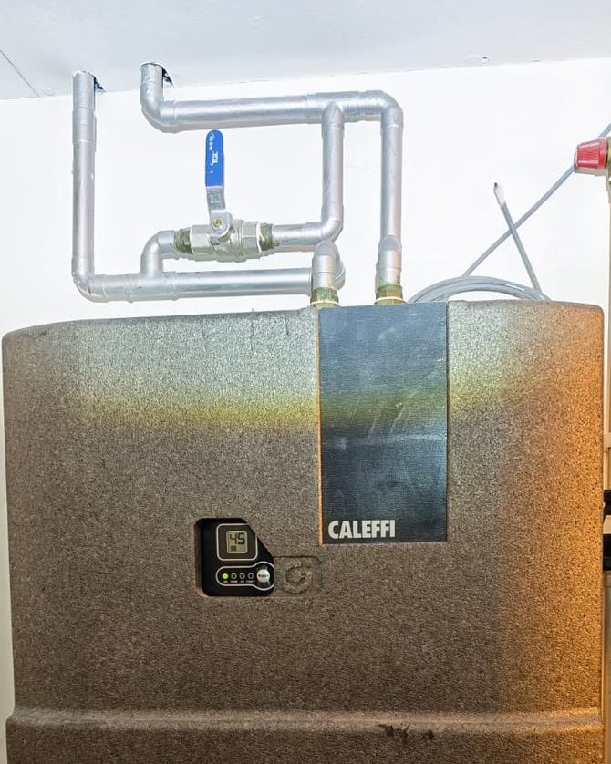 Wall-mounted Caleffi heat interface unit in a flat cupboard, with galvanised pipework and a controller showing its set point