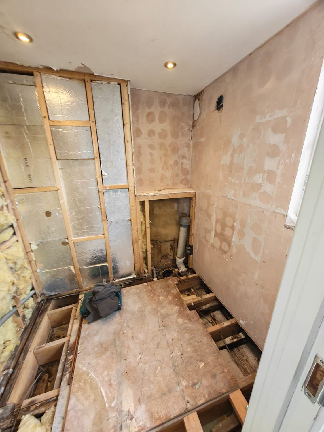 Shower room stripped back: the stud wall opened to foil-faced board, floorboards lifted and the waste pipework exposed